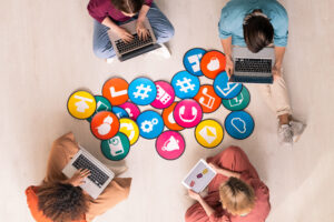 Top view of four young friendly people in casualwear sitting by multi-color papers with icons on the floor and using mobile gadgets